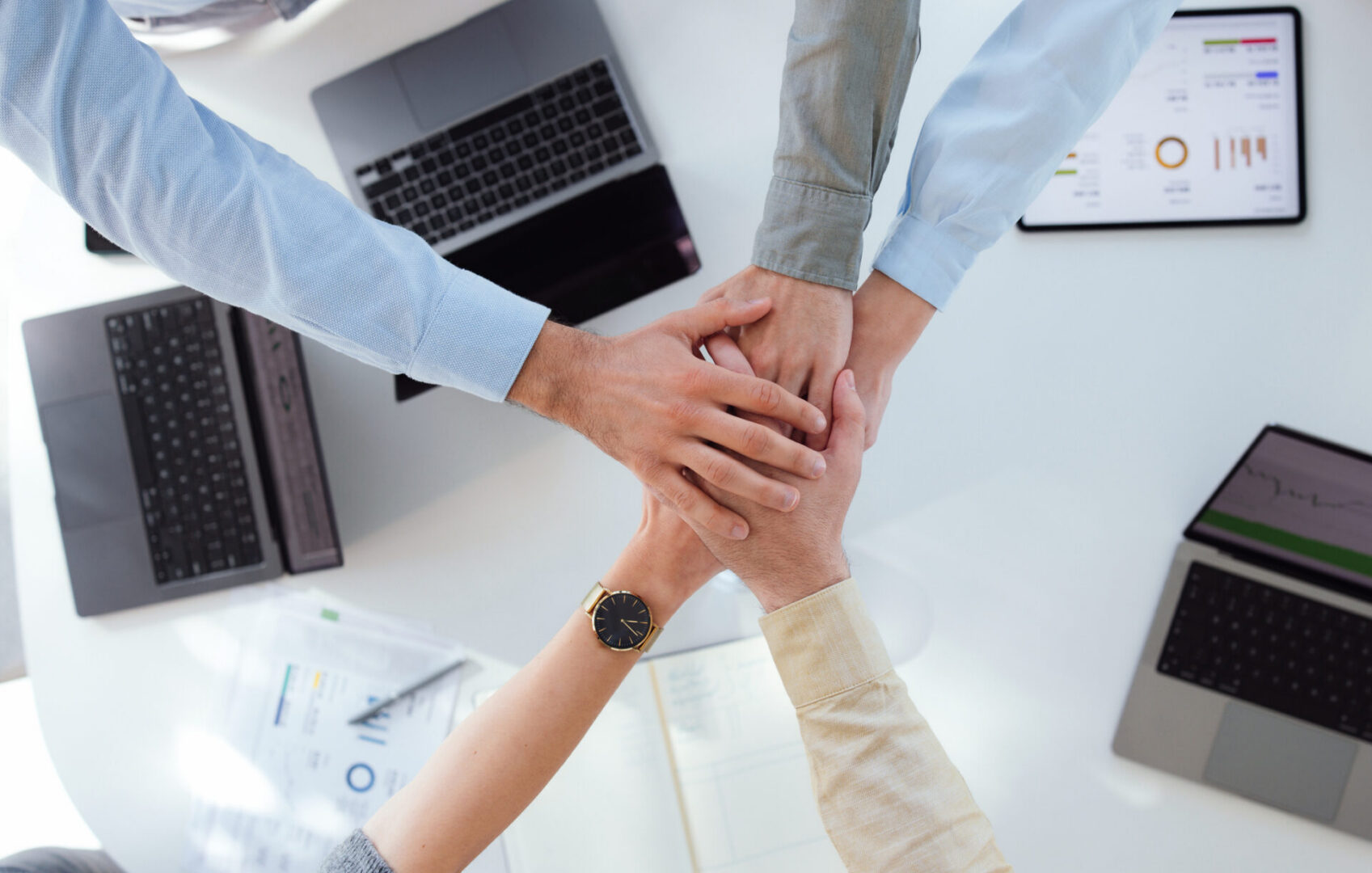 Colleagues join hands in a show of teamwork and unity around a white tabletop with laptops and reports, conveying collaboration, trust, partnership and shared commitment in a modern office work environment.